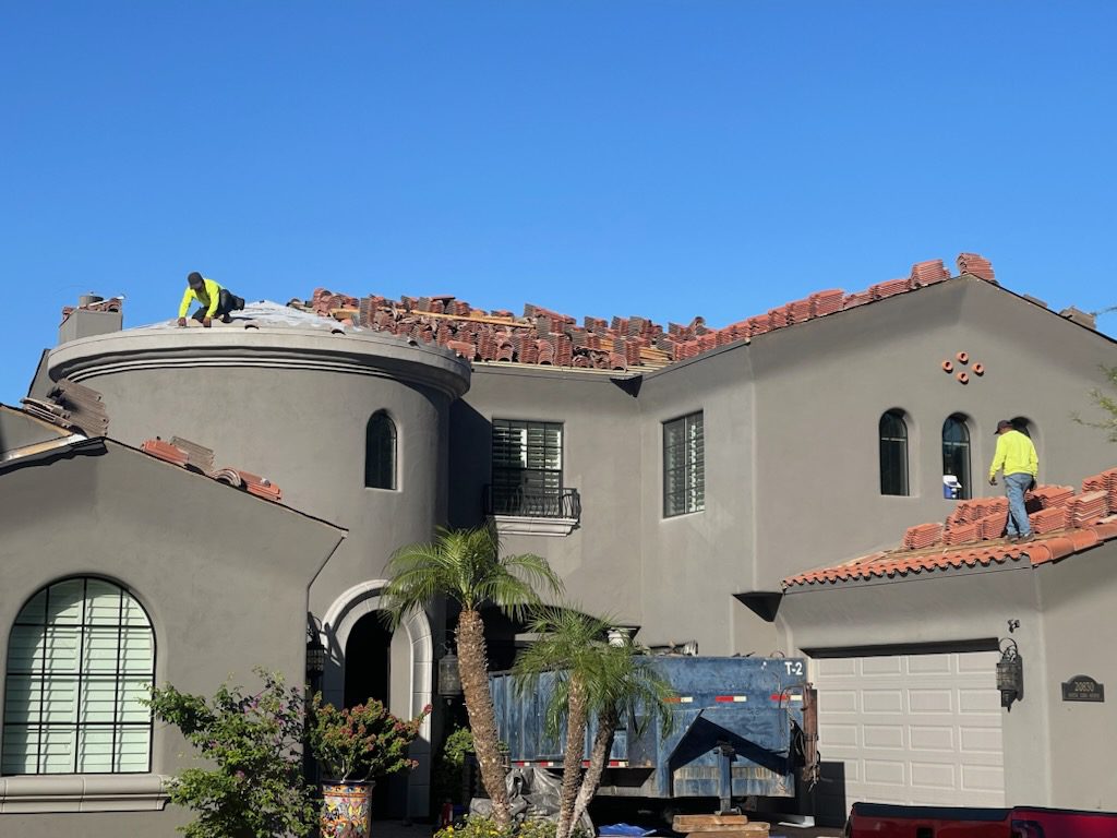 Workers repairing roof tiles on a large house under clear blue sky.