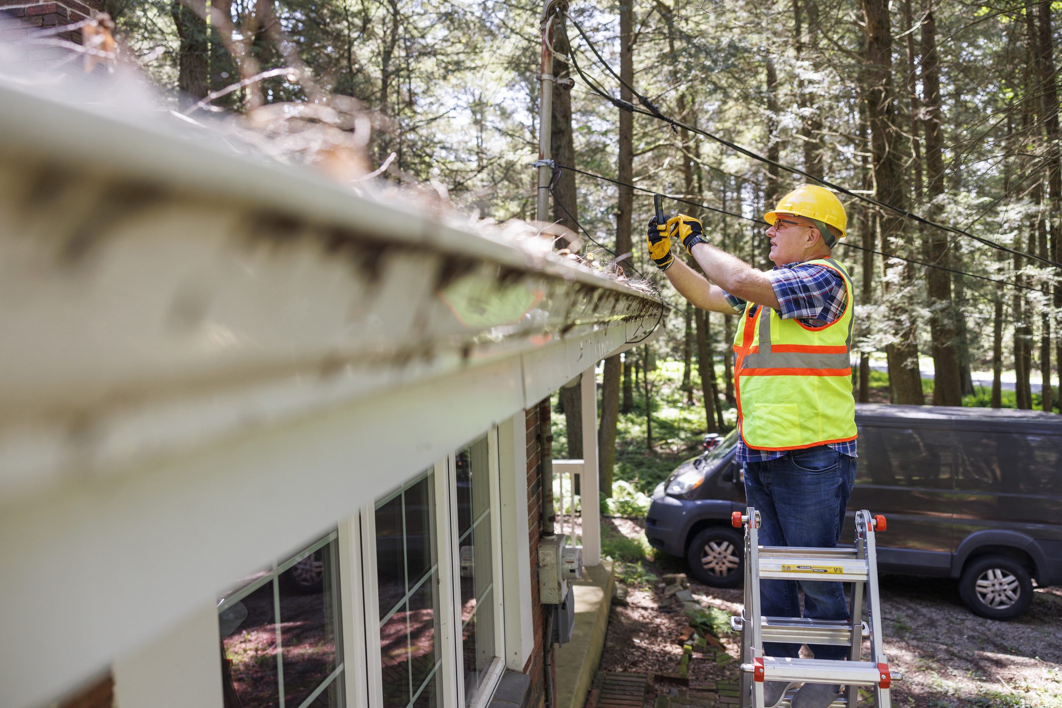 Worker cleaning gutters on a house using a leaf blower.