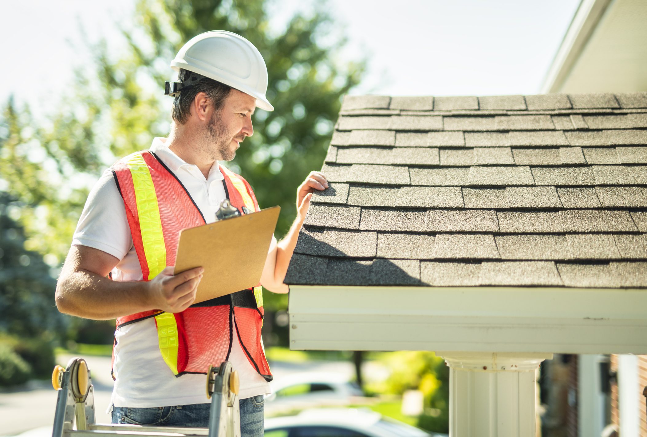 Construction worker inspecting roof shingles with clipboard in hand.